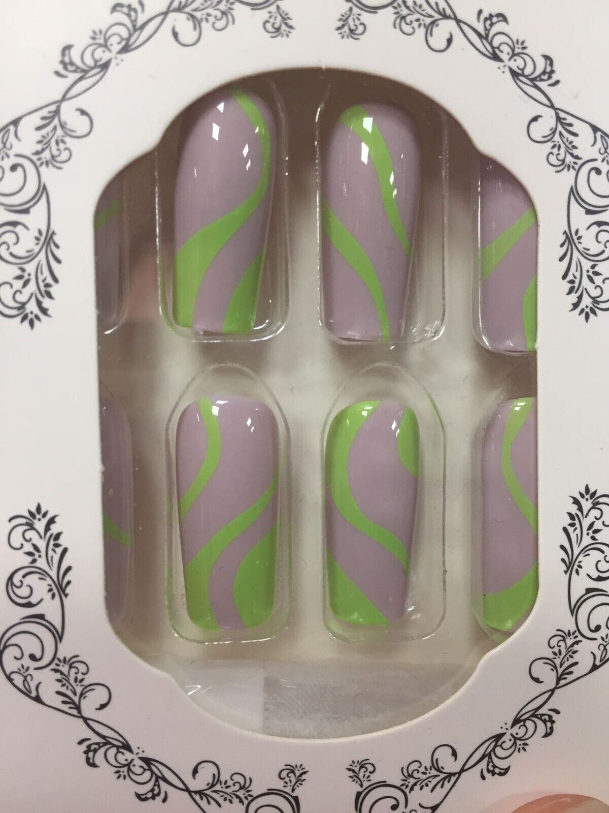A set of six press-on nails in a decorative white tray with black floral scrollwork. The nails have a light lavender-purple base with a bright lime green wavy stripe pattern.
