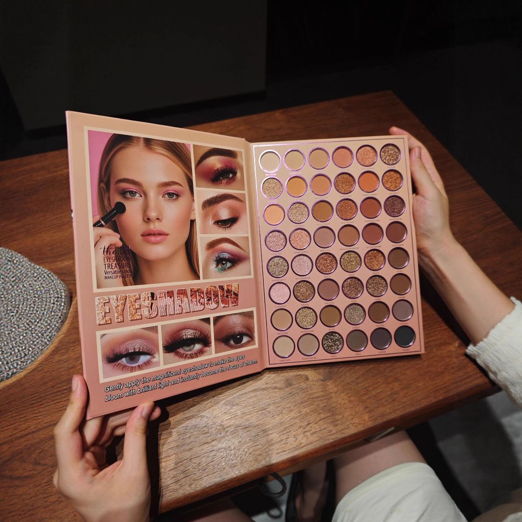 A person holds open a large eyeshadow palette on a wooden table. The palette cover shows a model wearing dramatic eye makeup, and the open section contains many round eyeshadow pans in warm neutral and pink tones.