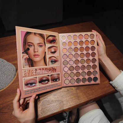 A person holds open a large eyeshadow palette on a wooden table. The palette cover shows a model wearing dramatic eye makeup, and the open section contains many round eyeshadow pans in warm neutral and pink tones.