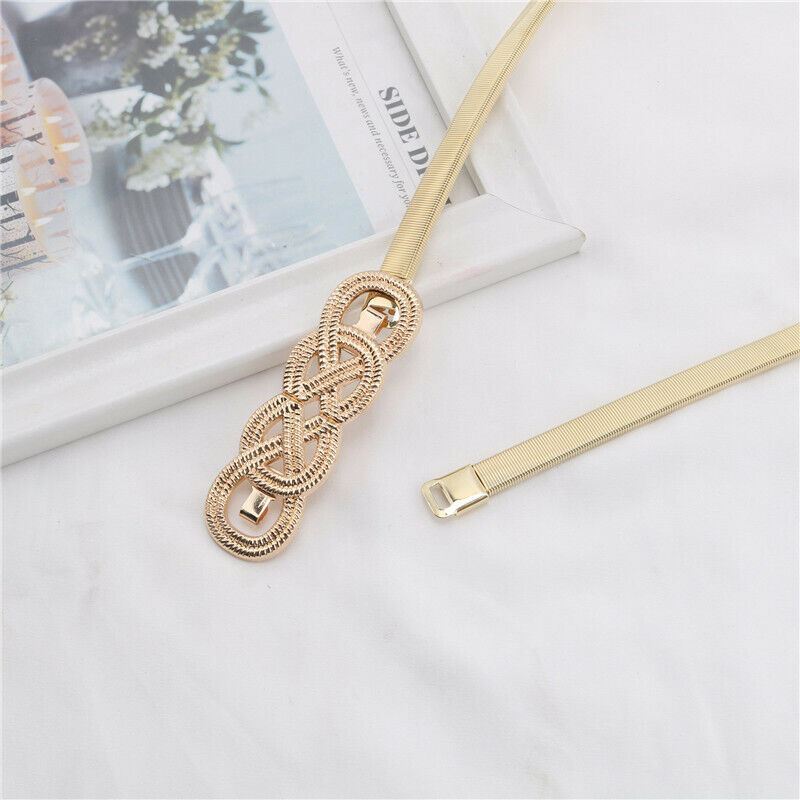 A gold-toned metal belt with a decorative braided knot center and a matching mesh strap, laid on a white fabric surface next to a white book or magazine.