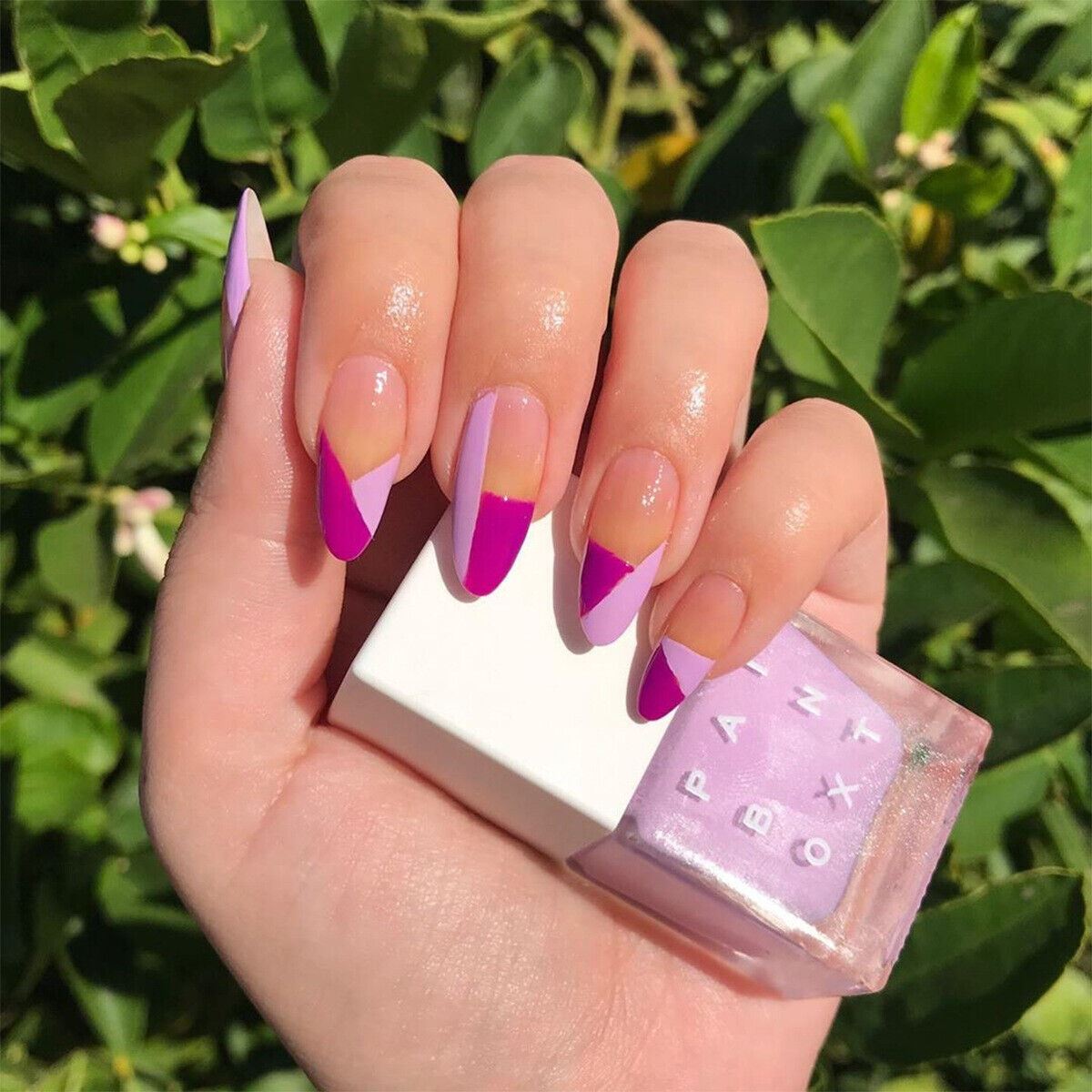 Hand with pink and purple gradient nails holding a nail polish bottle against a green leafy background