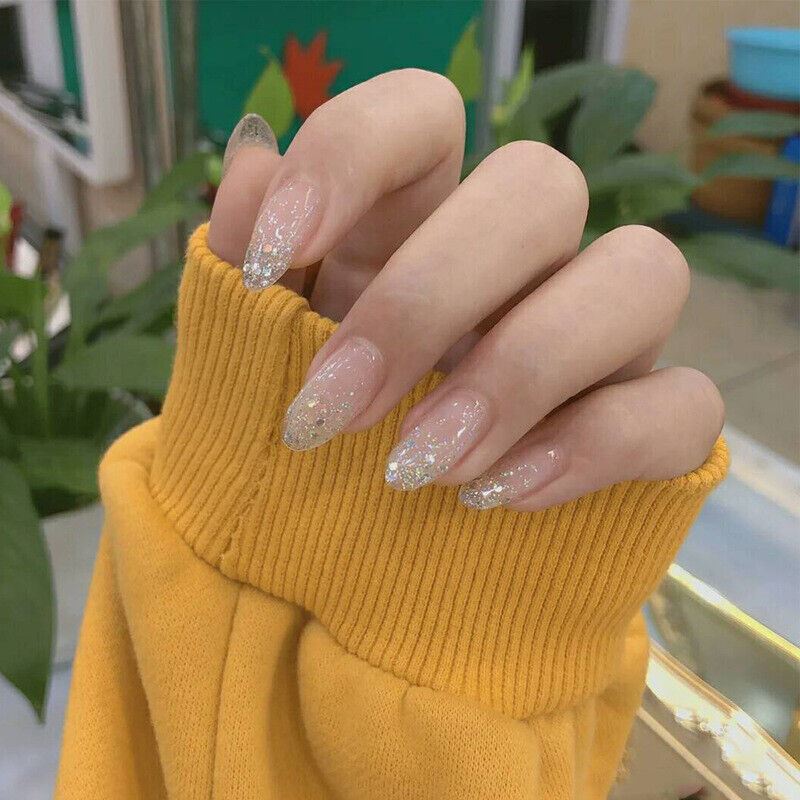 A close-up of a hand wearing long, almond-shaped press-on nails with a sheer nude base and fine silver glitter, styled with an oversized yellow knit sweater cuff.