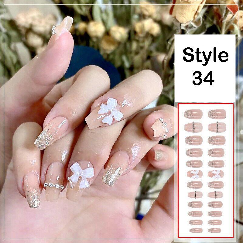 A hand displaying press-on nails in style 34, featuring a nude pink base with silver glitter tips, white bow and clover designs, and rhinestone accents, alongside a product sheet showing the nail shapes.