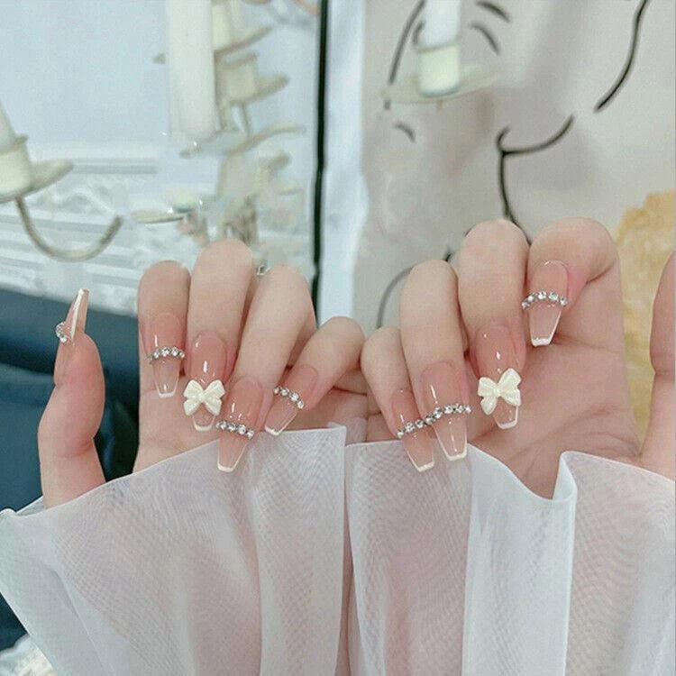 A pair of hands displaying coffin-shaped press-on nails with a nude pink and white ombre gradient, decorated with small white bows, clear rhinestones, and delicate pearl chains.