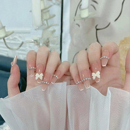 A pair of hands displaying coffin-shaped press-on nails with a nude pink and white ombre gradient, decorated with small white bows, clear rhinestones, and delicate pearl chains.