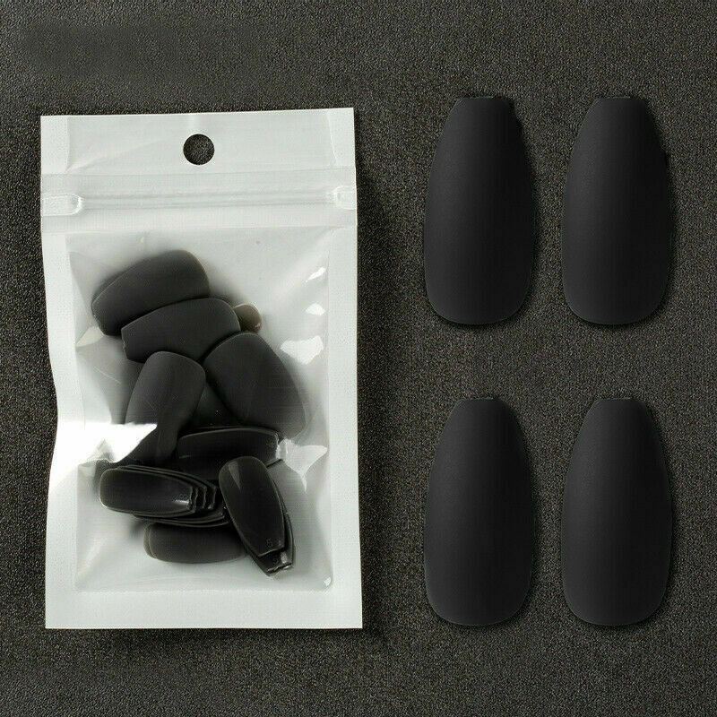 Four matte black coffin-shaped press-on nails displayed next to their clear plastic resealable pouch on a dark textured background.