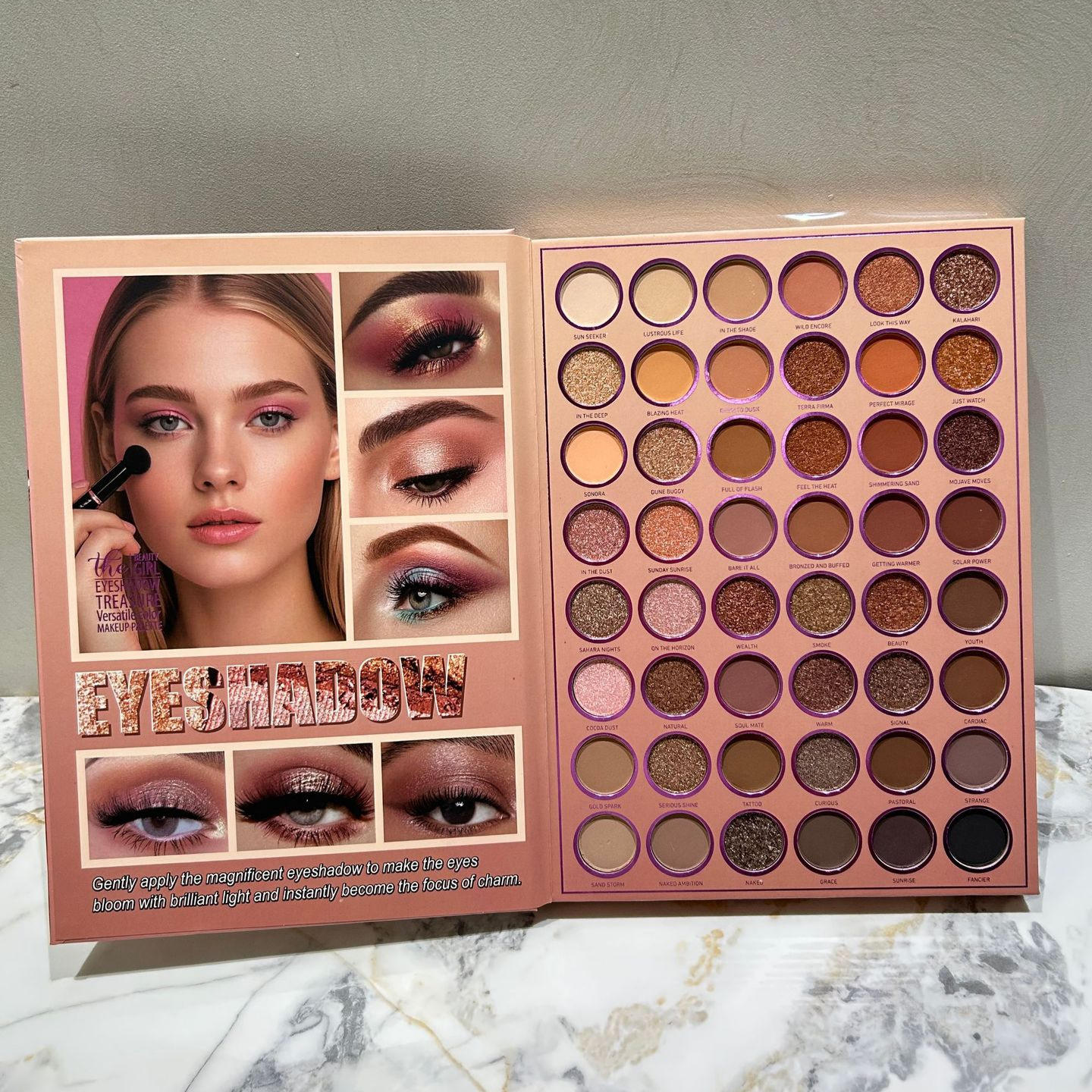 An open eyeshadow palette displayed on a marble surface against a gray wall. The left page shows a model wearing the eyeshadow makeup with before-and-after eye makeup panels and the text 'EYESHADOW'.