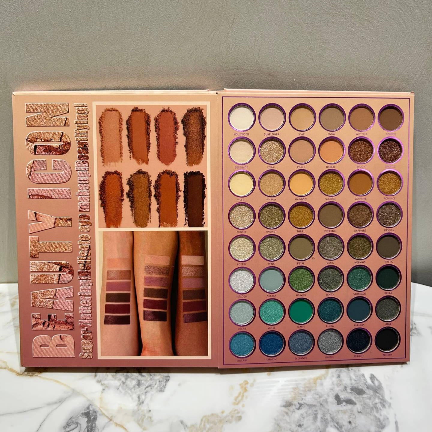 An open Beauty Icon eyeshadow palette displayed on a marble surface against a gray wall. The palette features a rose gold-toned cover with 'BEAUTY ICON' printed vertically, alongside swatches of lip and eye colors, and 36 round eyeshadow pans in neutral, earthy, and cool tones.