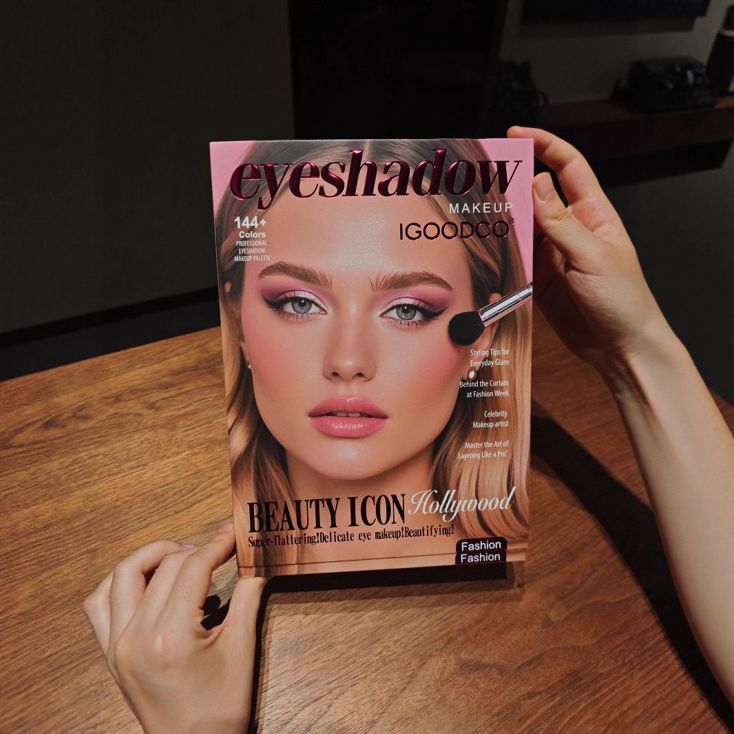 A person holds an eyeshadow makeup book titled 'eyeshadow MAKEUP iGOODOO' featuring a model's face with pink eyeshadow on a wooden table.
