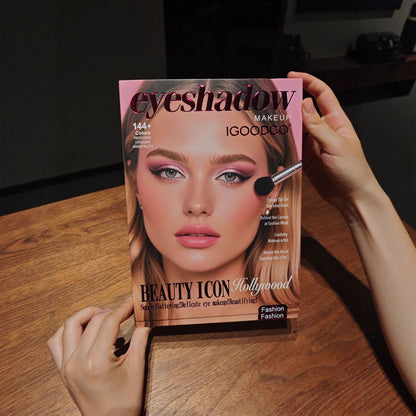 A person holds an eyeshadow makeup book titled 'eyeshadow MAKEUP iGOODOO' featuring a model's face with pink eyeshadow on a wooden table.