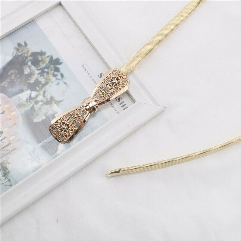 A gold-tone snake chain necklace with a decorative filigree bow-shaped pendant, styled on a white marble surface next to a white picture frame.
