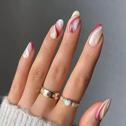 A close-up of a hand with almond-shaped nails decorated in a pink, white, and pale yellow marble swirl pattern. The hand is wearing two gold rings, one with heart details, against a neutral gray background.