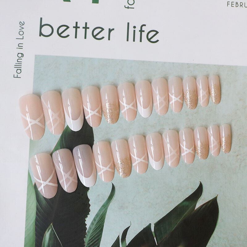 A set of press-on nails arranged on a magazine page with tropical leaves. The nails have a glossy nude-pink base with white geometric starburst patterns on the front and gold glitter on several tips.