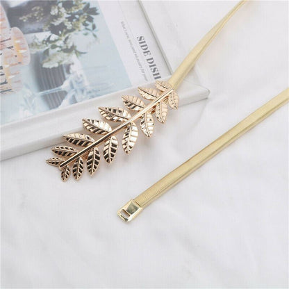 A gold-tone metal belt featuring a detailed leaf branch motif on one end and a slim mesh strap on the other, displayed on a white fabric surface with a magazine in the background.