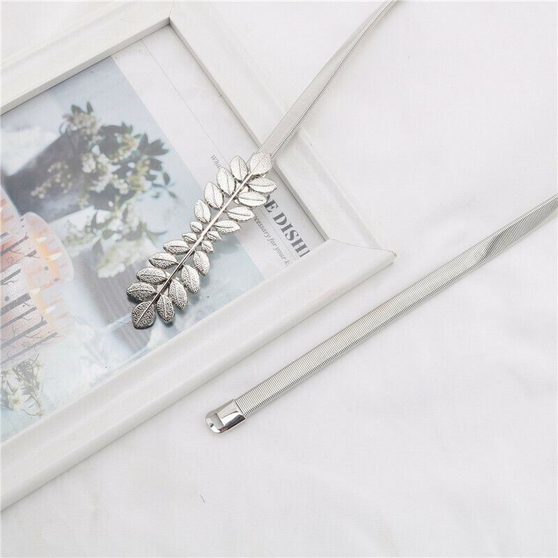 A silver metallic leaf-shaped hair claw clip attached to a slim silver elastic band, displayed on a white fabric surface next to a white picture frame.