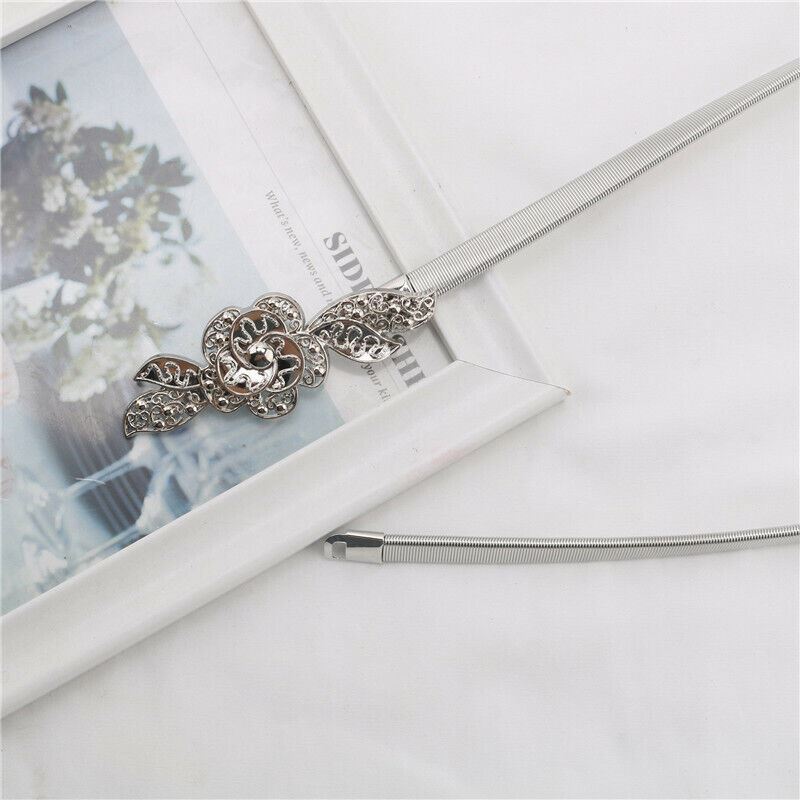 A silver-toned metal necklace with a detailed filigree flower pendant and a textured snake chain, styled on a white surface next to a white picture frame.