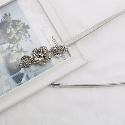 A silver-toned metal necklace with a detailed filigree flower pendant and a textured snake chain, styled on a white surface next to a white picture frame.