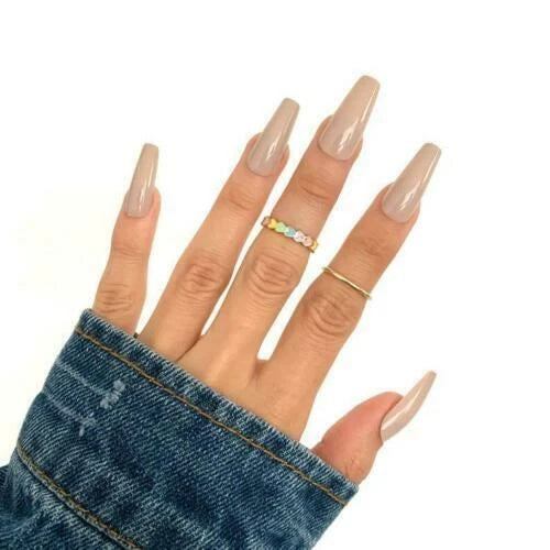 A close-up of a hand with long, glossy nude-colored almond-shaped nails, wearing two delicate rings on the middle and ring fingers. One ring features small pastel-colored stones, and the other is a thin gold band.