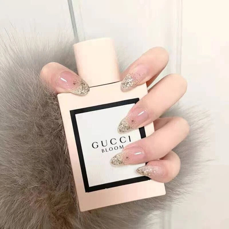 A hand with long, glossy nude nails decorated with silver glitter holds a bottle of Gucci Bloom perfume. The perfume bottle is a pale pink with a white label featuring a black border and the text 'GUCCI BLOOM'.