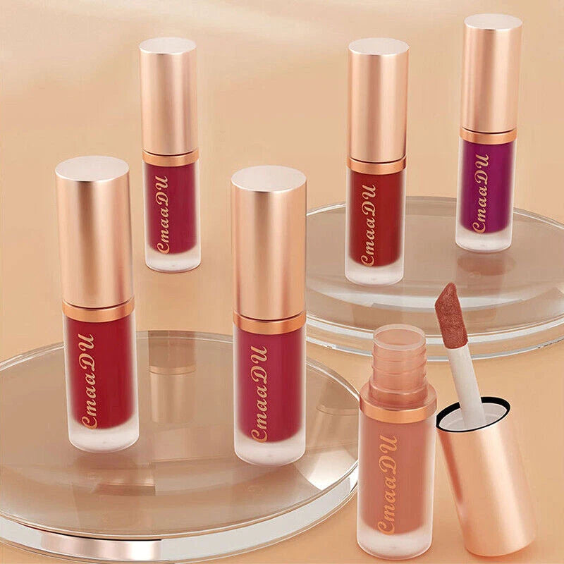 Five tubes of EmmaDU liquid lipstick in various shades, including deep berry, plum, nude, and mauve, arranged on clear glass display stands against a warm beige background. One tube is open with the applicator removed, showing the brand name 'EmmaDU' printed vertically on the tube.