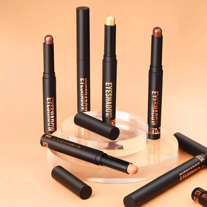 Five black cosmetic eyeshadow sticks with gold lettering are arranged on a clear acrylic stand against a warm beige background. One eyeshadow stick is shown uncapped with its peach-colored product visible.